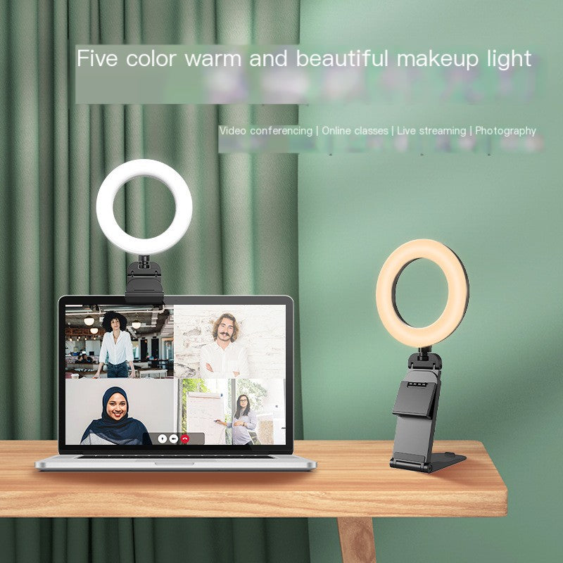 Two adjustable smartphone ring lights on stands, one emitting cool white light and the other warm yellow light, displayed on a wooden table with a laptop in the background against a green curtain backdrop.
